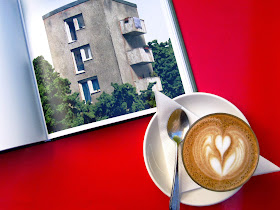 Aerial view of cafe table showing a cup of coffee and an open book with a picture of a model building in it.