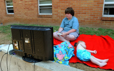 Overweight adolescent boy sits on ground OUTSIDE playing video game on a large television