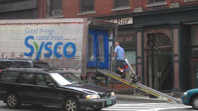 Silver Sysco truck unloading on the street with ramp down, blue and green logo with leaf and the tag line Good things come from Sysco