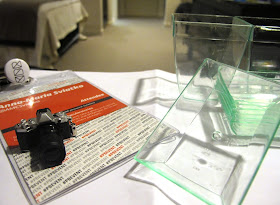 A conference lanyard on a desk in a hotel room. On it is a miniature camera, and next to it are a number of clear plastic containers which look like miniature planters and sinks.