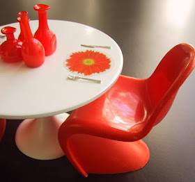 Modern dolls' house miniature Sarinnen table and orange Panton chair. On the table are some orange glass bottles and a place mat in the shape of a flower.
