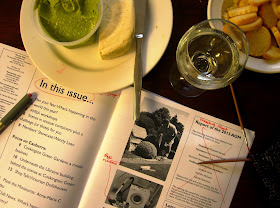 Aerial view of a coffee table, showing cheese and crackers and dip, a glass of wine, a marked-up copy of a magazine and some knitting in progress.