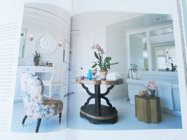 Bathroom in book by Kitchens and Baths by Michael S. Smith with a white stand alone tub, upholstered flower print arm chair, white vanity with gold drawer pulls, and a table