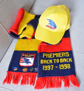 A Crows scarf with a yellow peaked cap resting on top.  The cap has a Crows logo on the front.  The scarf has horizontal stripes in navy, red and yellow with red fringing at each end.  One end of the scarf has an Adelaide Crows patch stitched onto it with membership badges pinned along the bottom edge.  The other end of the double-sided scarf has the words "Premiers back to back 1997–1998" machine-knitted in fair isle. 