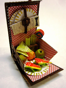 A miniature picnic hamper with red checked lining, holding two glasses, a lollipop, two smoked salmon and lettuce sandwiches, two apples and an orange.