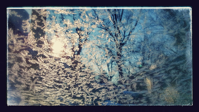 Jagged frost on a window reflects rising sunlight with trees and a small house in the background.