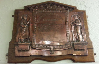 A bronze or brass decorated plaque on a wooden background, mounted high up on a wall, so difficult to get a square picture.