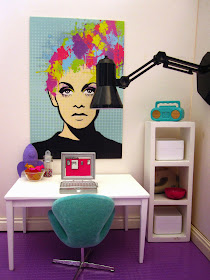 Modern dolls house miniature scene of a study with white walls and purple tiled flooring. A teal swan chairs sits in front of a white table with a laptop on it. A white shelving unitl is to the right and a large place lamp is attached to the right-hand wall.