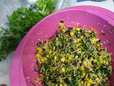 A pink bowl filled with a mixture of corn, black beans, shredded cheese, and chopped greens. Fresh cilantro is seen to the side, partially in a transparent plastic bag.