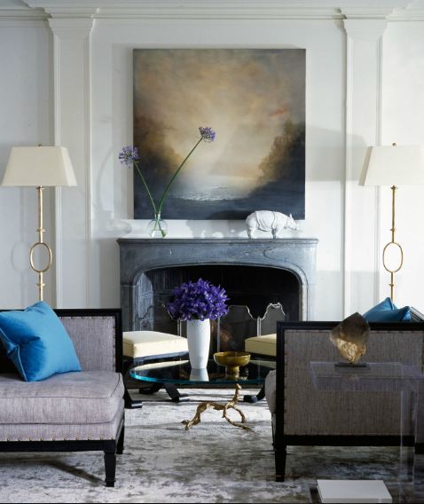 Living room with grey upholstered seating with nail head trim and turquoise accent pillows, a dark grey fireplace with tall brass lamps on each side and white panel molded walls