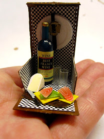 A miniature picnic hamper with black and white checked lining, holding a bottle of wine, two glasses, two smoked salmon sandwiches and a wedge of camenbert, displayed on an outstretched hand.