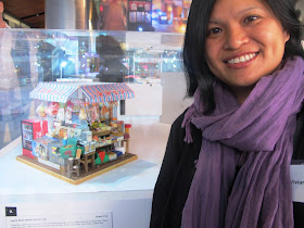 A woman standing next to a miniature model of a traditional Hong Kong Joyful Store.