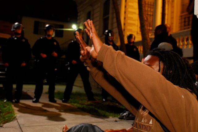 Fully-geared riot cops surround a young Black protestor sitting with his hands in the air.