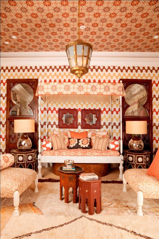 Bedroom by Martyn Lawrence Bullard with red swiggly wallpaper on the walls and a wallpapered ceiling, white canopy bed, Moroccan mirrors and nightstands and two armchairs