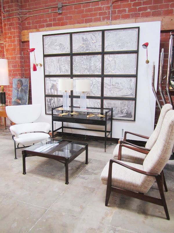 Room with brick wall with an Old map cut up and framed in sections behind a glass viewing table, three armchairs and a coffee table with a glass table top and iron legs