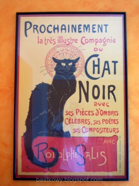 Fotografia przedstawiająca plakat "Czarny Kot" zawieszony na ścianie w czarnej ramie, nadającej wnętrzu artystycznego charakteru