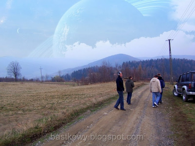 Grupa ludzi spacerująca po wiejskiej drodze z dodanym w Photoshopie obrazem planety Saturn na niebie, co nadaje zdjęciu nierealistyczny, ale intrygujący wymiar.