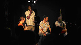 Tostan's Seydou Niang (standing) presents about Tostan France during the concert. Volker (seated, left), Seydou (standing, back), Percussionist Joe Quitzke (seated, middle), and Cissokho (seated, right).