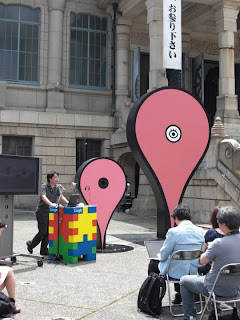 築地本願寺に設置されたGoogle マップピンと隣でスピーチ中の方を示す画像。