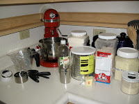 Counter covered in all the ingredients needed to make the whole wheat bread. Includes sugar and a red kitchenaid mixer.