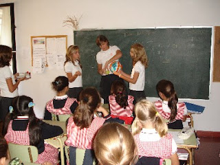 Fotografía de sala de clases, frente a la pizarra, una profesora y dos estudiantes desarrollan una actividad con una pelota