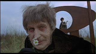 Dr. Phibes em primeiro plano, seu rosto em close, segura duas margaridas próxima ao nariz e sorri. Atrás dele uma estrutura abstrata de madeira. Ao fundo, uma jovem desfocada com chapéu preto em pé em um gramado alto, e o céu azul claro.