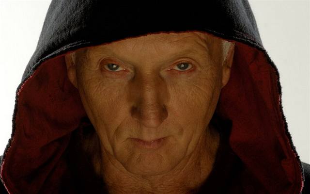 Close no rosto de Jigsaw (Tobin Bell): um homem branco de meia idade usando um capuz preto com forro vermelho. Ele olha para a câmera diretamente e com olhar morteiro, mas desafiador; sua boca ameaça um sorriso.