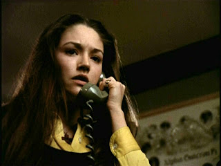 Uma jovem branca, de cabelo liso, longo e preto, segura ao ouvido o fone de um telefone verde oliva com fio. Sua expressão é de preocupação.