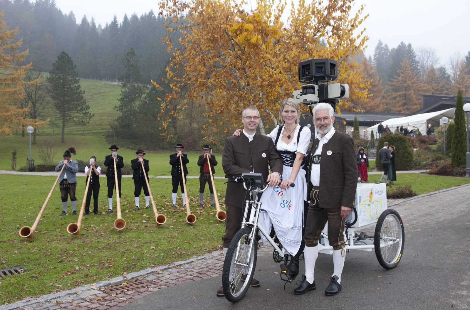 Ein Foto zeigt drei Personen auf einem Fahrrad mit Kameraaufbau. Im Hintergrund stehen Männer mit Albhörnern.