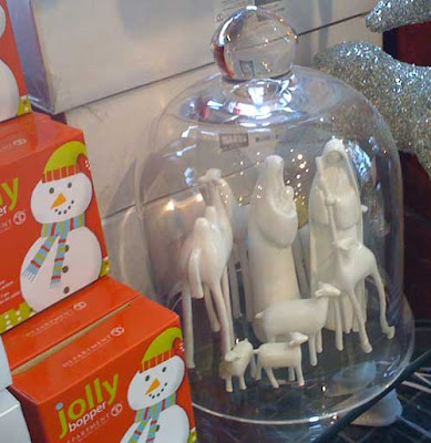 Photo of modernist, all-white ceramic figurines of Mary (holding Jesus), Joseph, a camel, and some sheep inside a clear glass bell jar