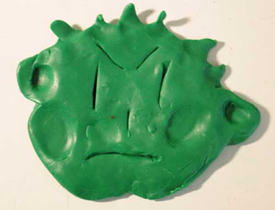 Flat green face made of clay, kind of like a kewpie doll