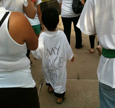 A four year old boy walks with a long white t-shirt almost to his ankles. Hand lettered on the back it says MY UNCLE with a sad, crying face below