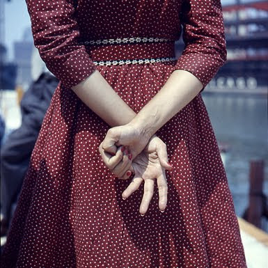 Young woman in dark red dress with white dots, shot from the back, no head shown, her hands clasped and her fingers making a sign of some sort