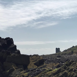 A picture of a ruined tower standing in the distance with rocks in the foreground.  This is Seafield Tower as seen from rocks by the Forth.  Photo by Kevin Nosferatu for the Skulferatu Project.