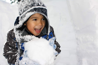 young boy in a beanie and ski jacket holding a giant snow ball. His mouth is open with excitement and a smile