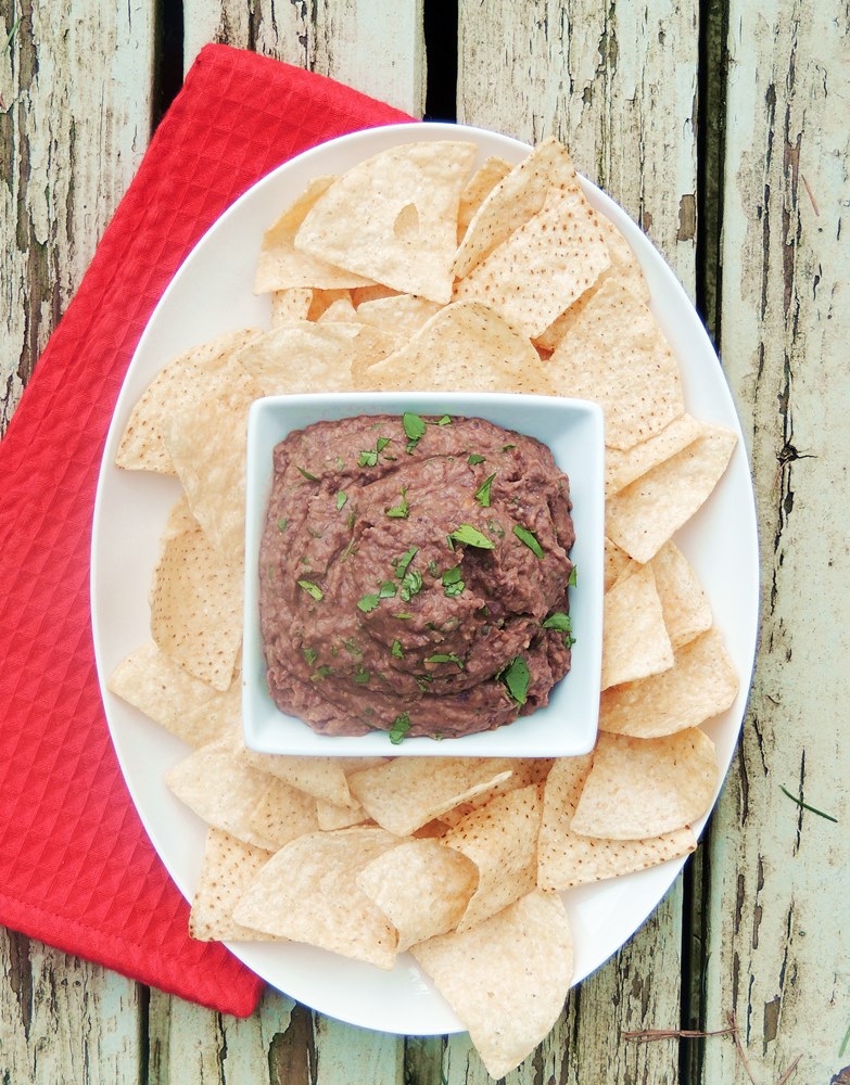 Bean dip in a square while bowl on a white platter with tortilla chips. Red towel and wooden background.