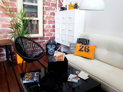 One twelfth scale modern miniature loft scene with brick exposed walls and wooden floors. To the right is a seating area with a futon sofa, trolley coffee table and outdoor chairs. On the coffee table is an open storage box and a selection of papers, photos and postcards. Next to it is a small rubbish bin.