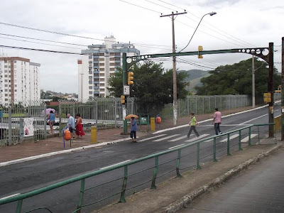 A foto apresenta um dia com nuvens pesadas e um leve chuvisqueiro. Pedestres atravessam a avenida Bento Gonçalves sobre a faixa de pedestres, em Porto Alegre. Algumas pessoas estão com guarda-chuvas.