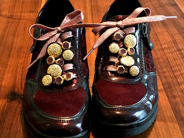 Napeilla tuunatut kengät edestä. Vaaleanpunaisissa kengännauhoissa isoja kultaisia nappeja ja pienempiä timantinmuotoisia nappeja, joissa on kultaiset reunat ja vaalean violetti keskus.