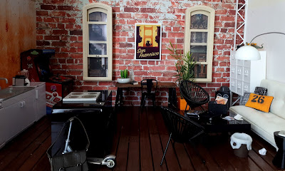 One twelfth scale modern miniature loft scene with brick exposed walls, long windows and wooden floors. To the left is a galley kitchen and an island bench. At the back between the windows is a trestle desk and to the right is a seating area with a futon sofa, trolley coffee table and outdoor chairs.