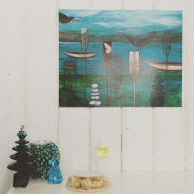 One-twelfth scale modern miniature Christmas scene on top of a sideboard with a beaded vase, tuny bead Christmas tree and glass angel next to a glass of sparking wine and a packet of chocolates. On the white paneled wall above is a picture in teals, blues and black.