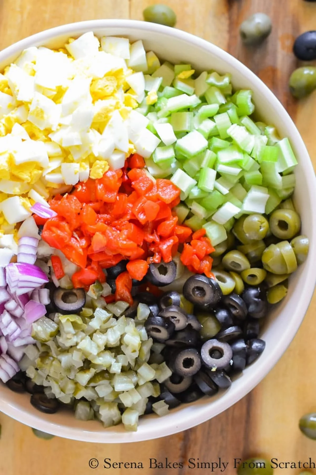 A large bowl filled with diced potato, sliced black olives, sliced green olives, diced egg, diced celery, diced onion, diced roasted red pepper, and diced spicy dill pickles.