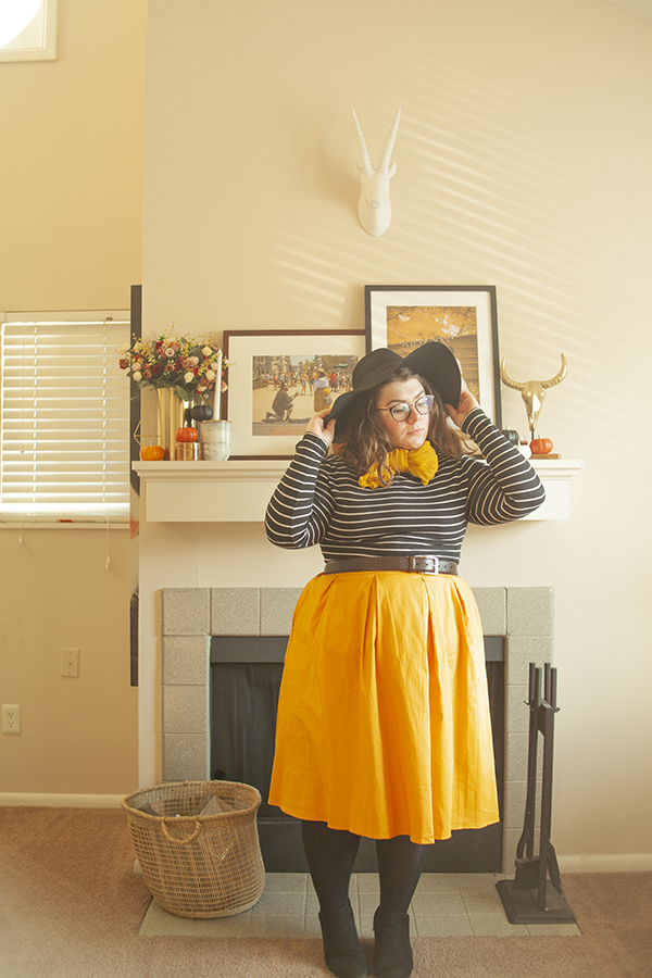 An outfit consisting of a wide brim black hat, striped mock neck shirt tucked into a yellow midi skirt, with tights and black ankle boots.