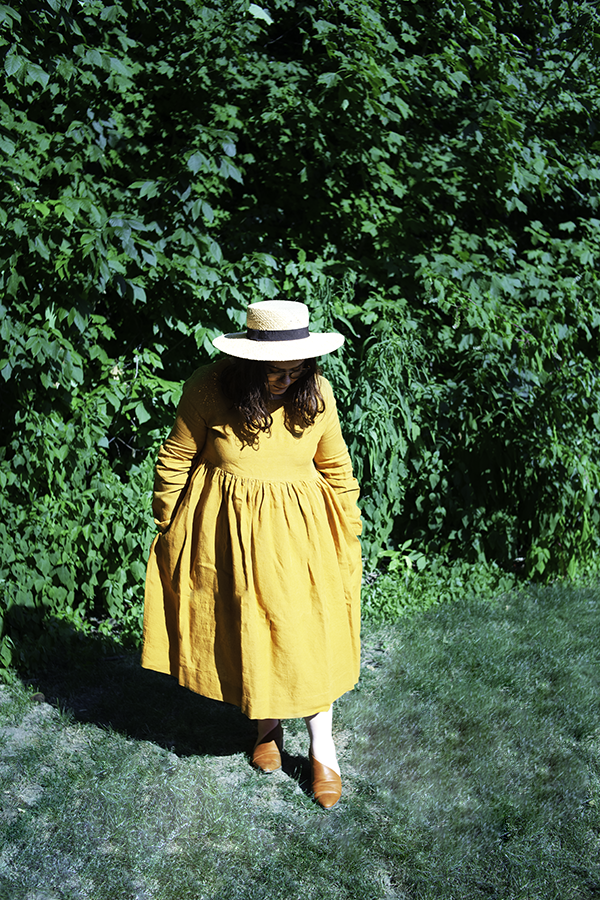 An outfit consisting of a straw boater hat, mustard yellow long sleeve linen smock dress, and cognac d'orsay flats.