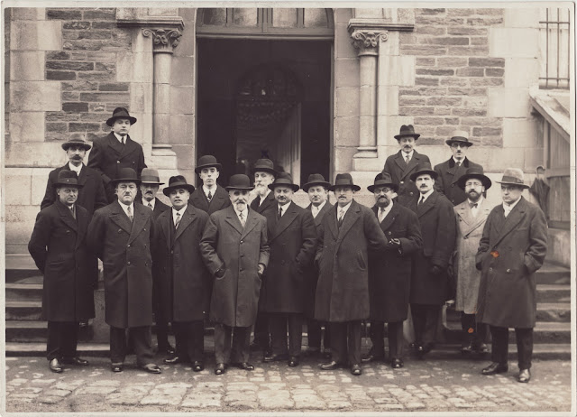 Le groupe des enseignants pose devant une porte ouverte. Les hommes ont tous des chapeaux et des pardessus.