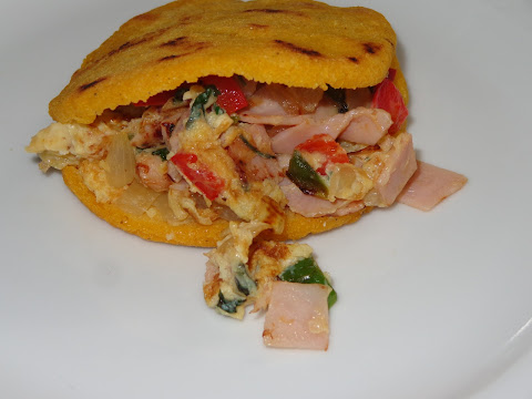 PORCIONES: 4  INGREDIENTES PARA HACER AREPAS 1 taza de harina de maíz precocinada  1 taza de agua ½ cucharadita de sal  METODO En un recipiente redondo mezclar bien la harina de maíz con el agua y sal, sin que queden grumos. Dejar la masa a reposar por 5 minutos. Con la masa formar 4 bolitas y aplastarlas dándole una forma redonda. Ponerlas sobre una sartén o plancha untada con un poquito de aceite. Poner la plancha o sartén a calentar y cocinar a fuego bajo las arepas por unos 8 minutos por lado. Deje dorar ambos lados. Cuando las arepas están tibias, abrirlas horizontalmente con un cuchillo, pero solamente hasta la mitad. Continuar abriendo hacia el fondo de la arepa pero dejando que los bordes queden juntos. Rellenar con lo que usted desee. En este caso las hemos rellenado con huevos revueltos y jamón.  HUEVOS REVUELTOS CON JAMON ½ cucharada de aceite 1 diente de ajo picado ¼ taza de cebolla cortada en cuadritos ¼ taza de pimiento rojo cortado en cuadritos ¼ taza de pimientos verdes cortados en cuadritos 1/2 cucharada de cilantro picado 2 lonjas de jamón 2 huevos batidos  METODO En una sartén calentar el aceite a fuego moderado y saltear el ajo con la cebolla, ajo, pimientos y cilantro. Agregar y calentar el jamón. Agregar y revolver los huevos dejando que coagulen. Rellenar las arepas con los huevos y jamón revuelto.   Las arepas es un platillo muy popular en Venezuela y Colombia aunque también lo hacen otros países. Consiste en una masa de maíz en forma redonda, por lo general cocinado a la plancha. Queda hecho con la forma de un pan. Se abre horizontalmente y se rellenan. Típicos rellenos son carne mechada, pollo con palta molida, huevos revueltos con jamón. Puede rellenar las arepas con lo que usted desee.