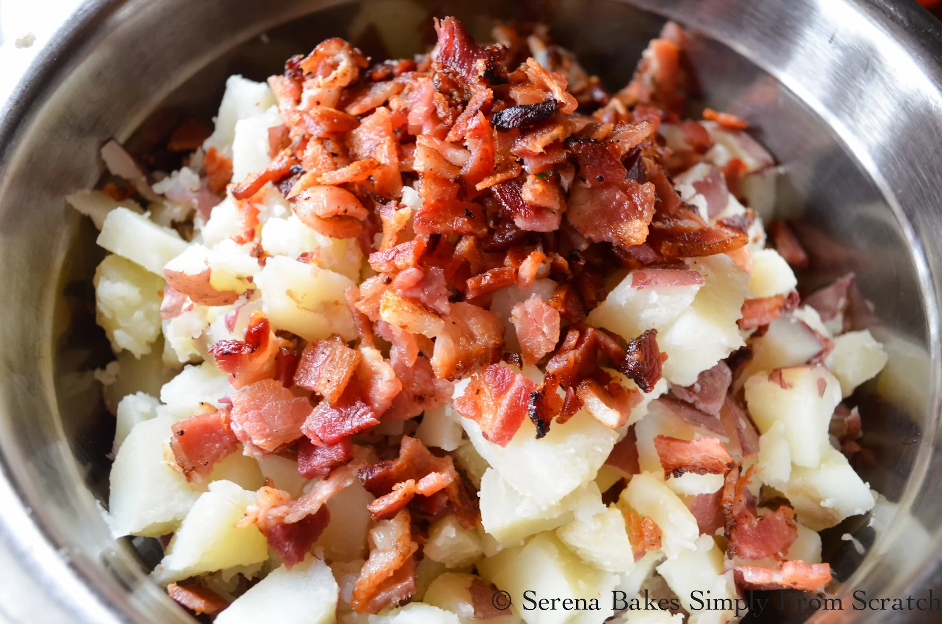 A large stainless steel bowl fully of cooked diced red potatoes with skins, and diced cooked bacon.