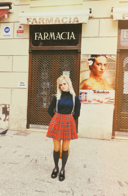 Alejandra Colomera vestida con jersey azul, falda roja cuadros escoceses, tirantes en color gris, calcetas gris y zapatos estilo colegial negro frente a farmacia antigua