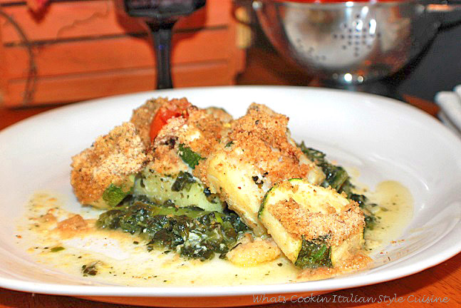 This is a baked cod fish on a white plate with a fall orange leaf basket in the background, a blue water glass and fork with napkin. The plate has a bed of spinach topped with baked cod fish with roasted vegetables on top along with bread crumbs toasted with Parmesan cheese and sliced tomatoes.