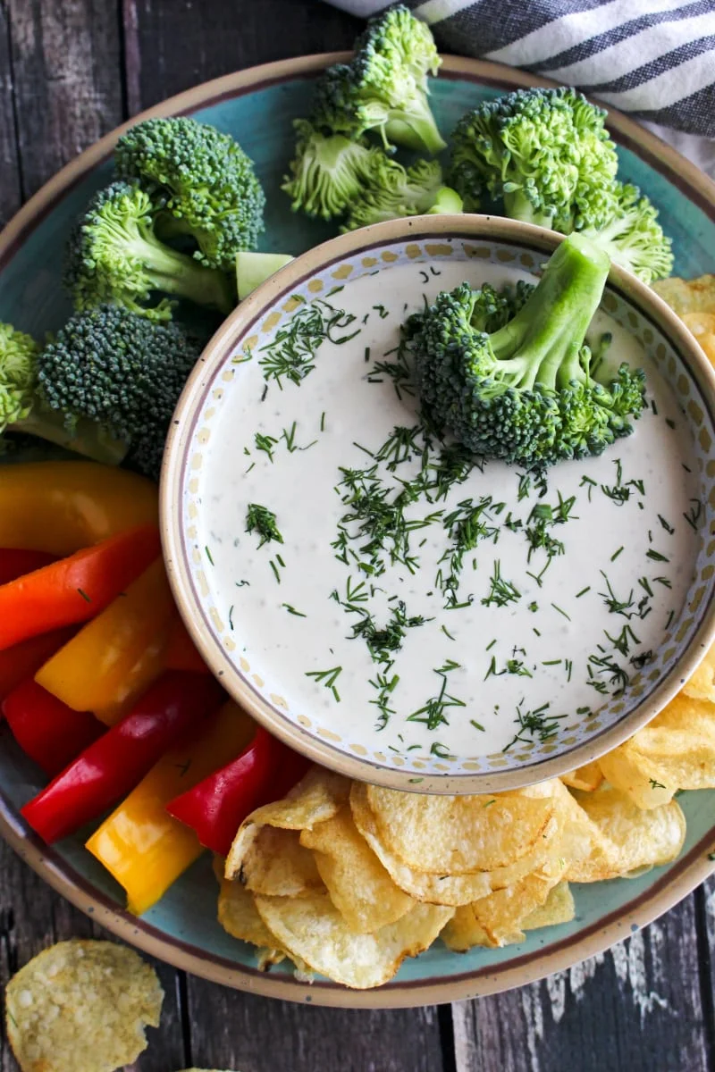 Close up top view of Dill Dip in a tan bowl on a blue plate surrounded by veggies and chips.